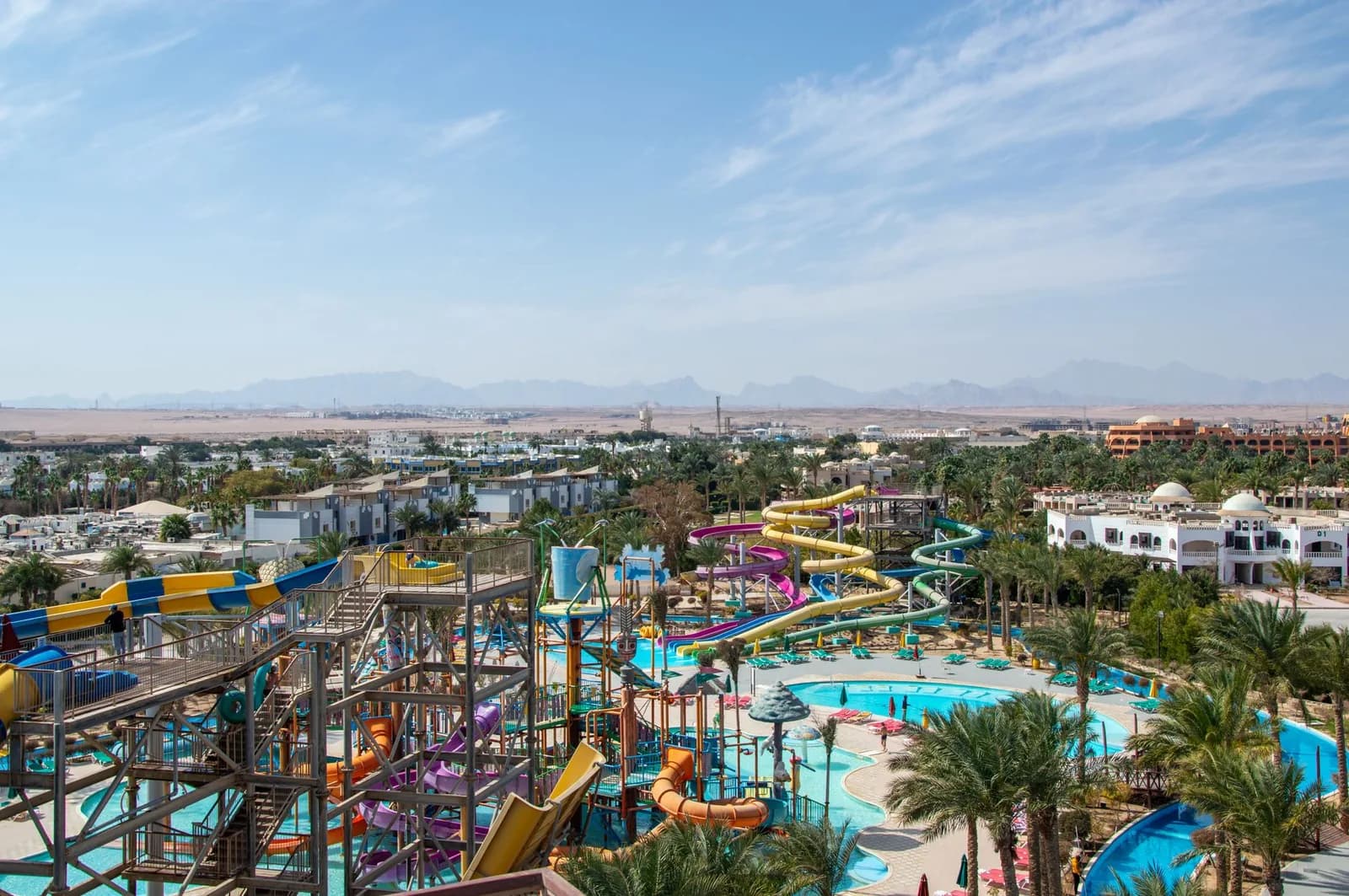Vue d'ensemble d'un grand parc aquatique de resort avec toboggans colorés, bassins et palmiers devant des montagnes arides