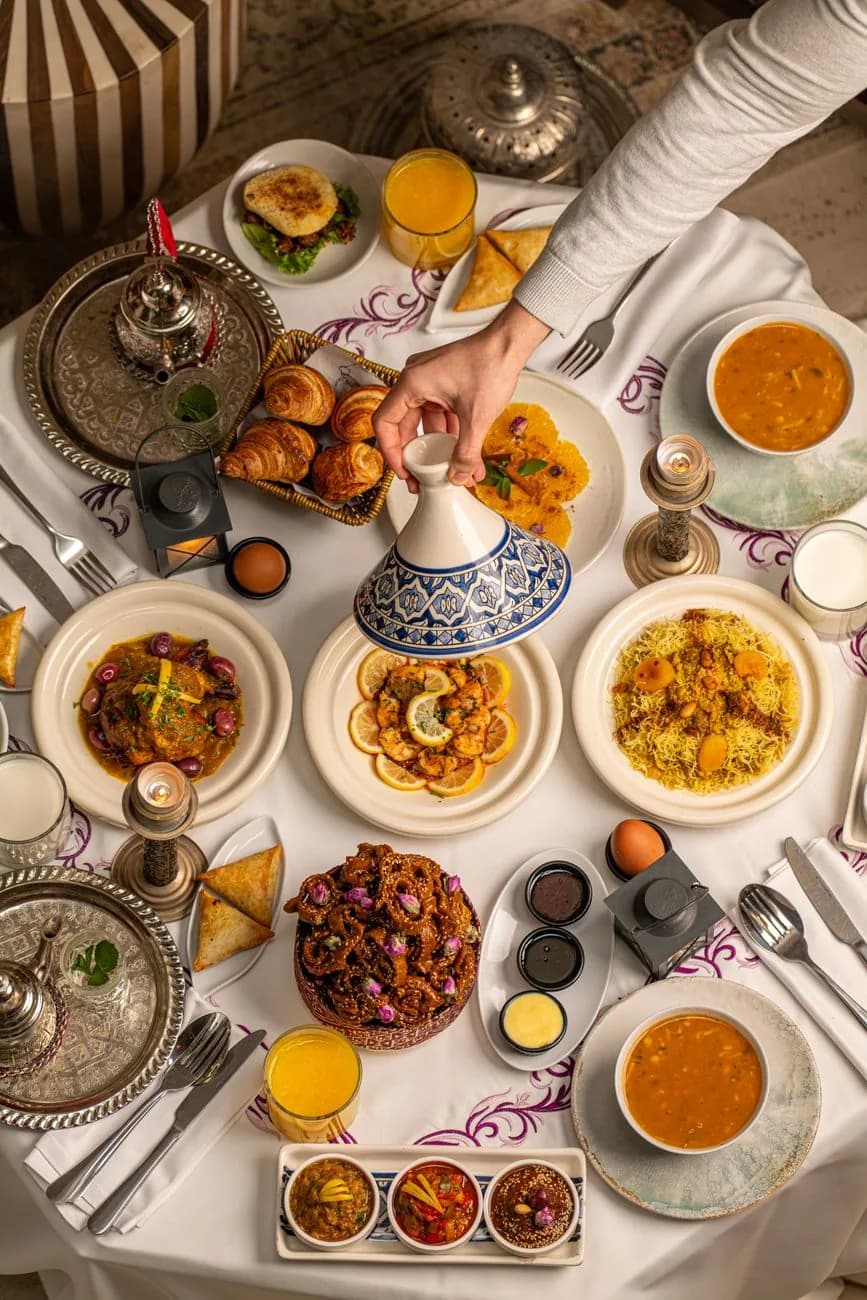 Table marocaine dressée à Marrakech, une main soulevant le couvercle d'un tajine au milieu des salades, du couscous et des pâtisseries