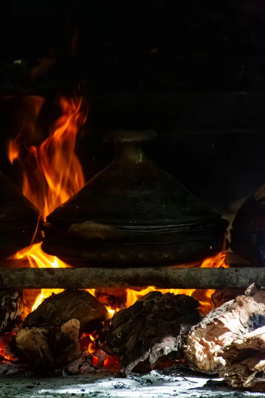 Tajine en terre cuite posé sur un feu de bois dans une cuisine de campagne marocaine, braises et flammes visibles