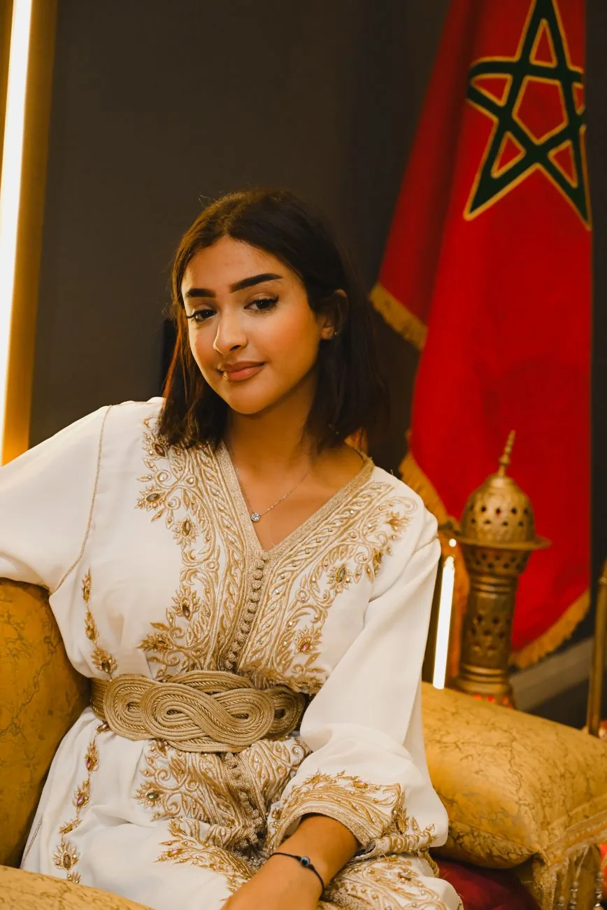 Jeune femme en caftan blanc brodé de fils dorés, devant un drapeau marocain et un brûle-encens en cuivre