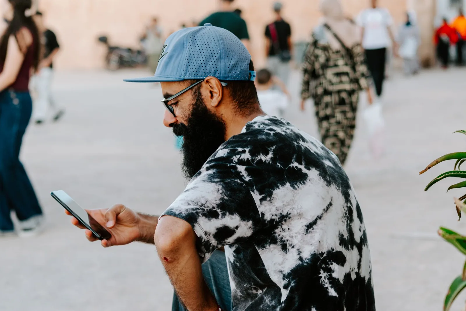 Homme à casquette qui consulte son smartphone sur une place passante, devant un mur de terre ocre
