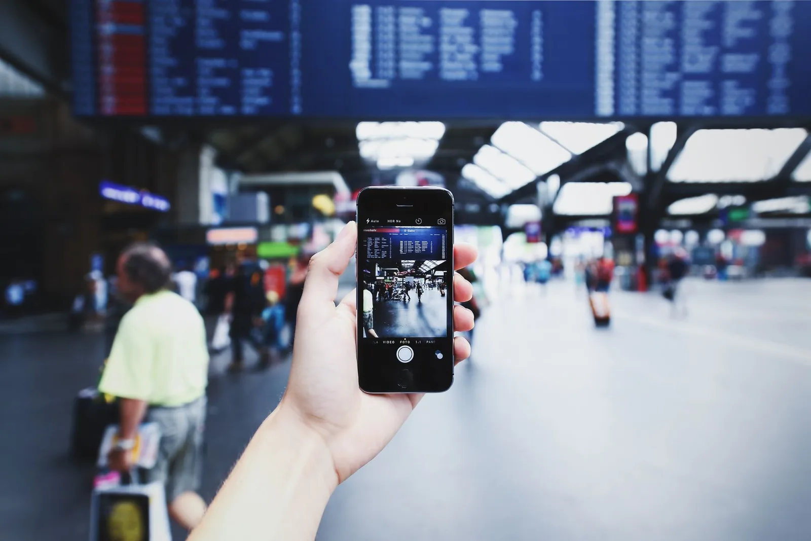 Main tenant un smartphone dans le grand hall d'une gare ou d'un aéroport, sous un panneau d'affichage des départs