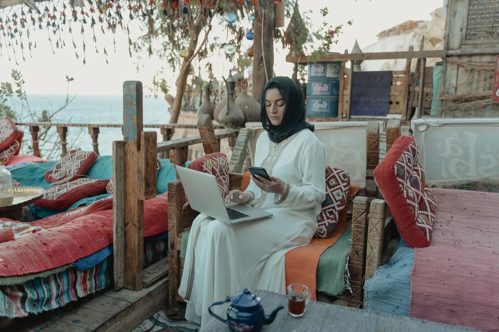 Femme assise sur des coussins d'une terrasse, avec un ordinateur portable, un smartphone et un verre de thé