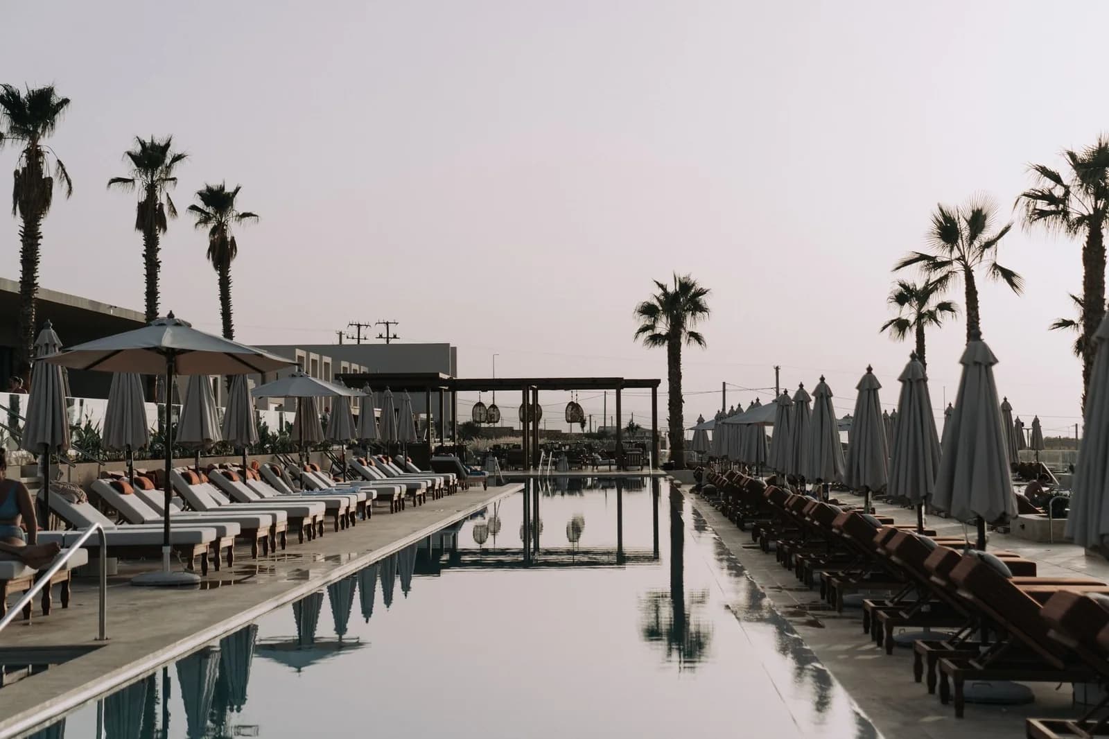 Longue piscine calme bordée de transats et de parasols fermés sous des palmiers, au crépuscule