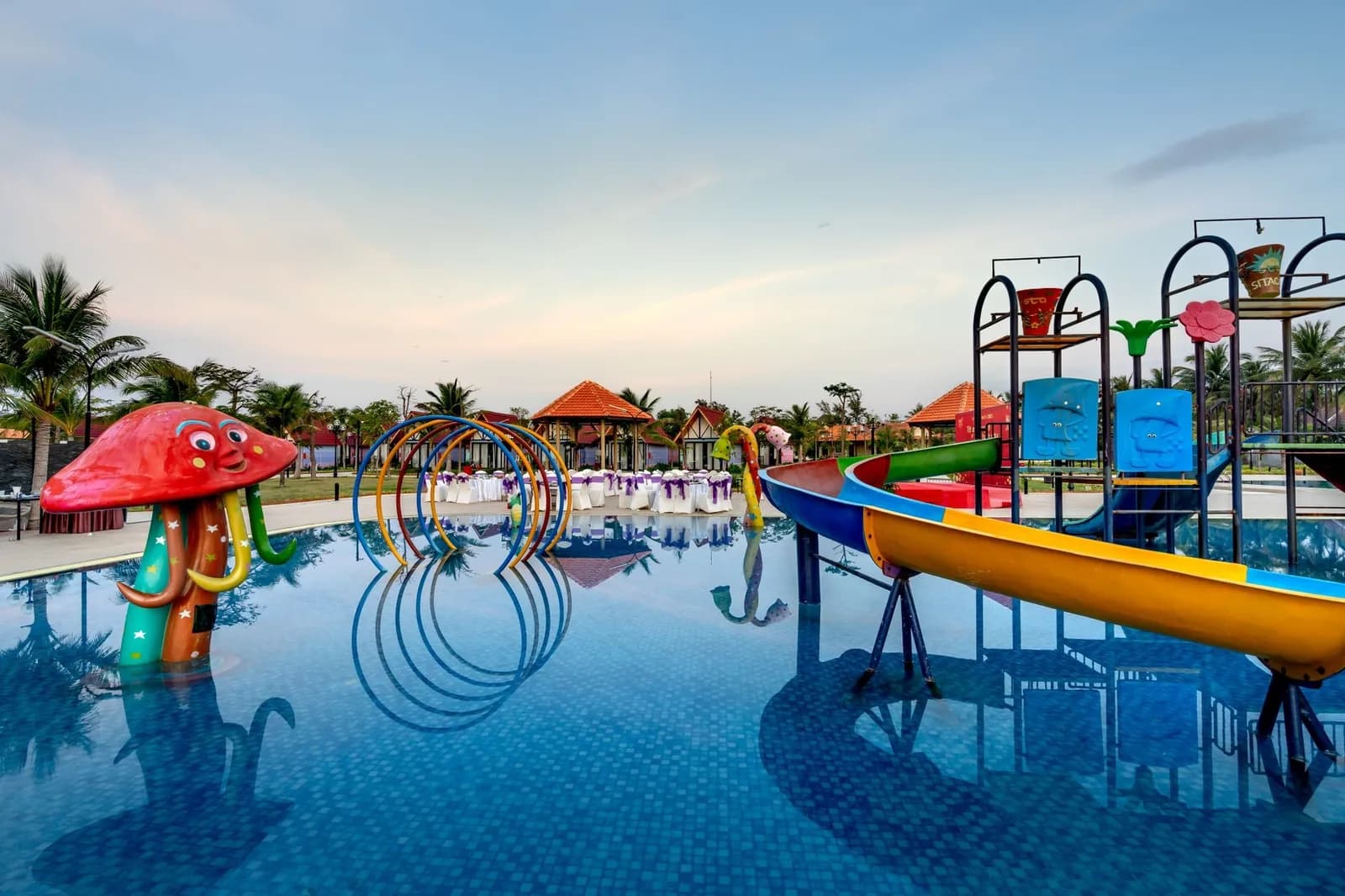 Aire aquatique d'un hôtel-club avec toboggans colorés, champignon jaillissant et arceaux d'eau au coucher du soleil