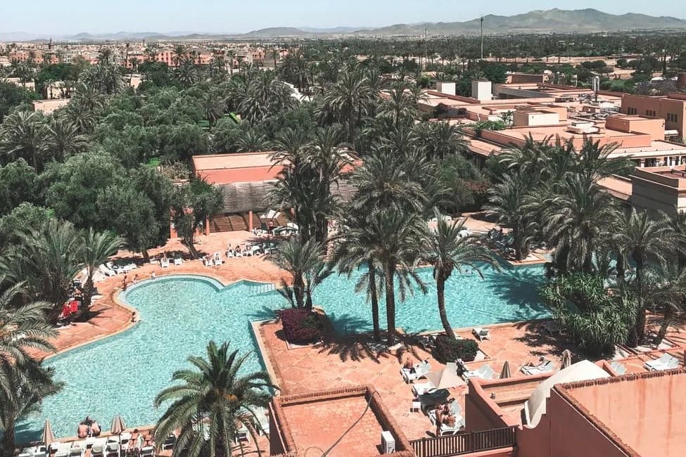 Vue aérienne d'un grand hôtel-club : piscine en forme libre bordée de palmiers, transats et toits ocre, montagnes au loin