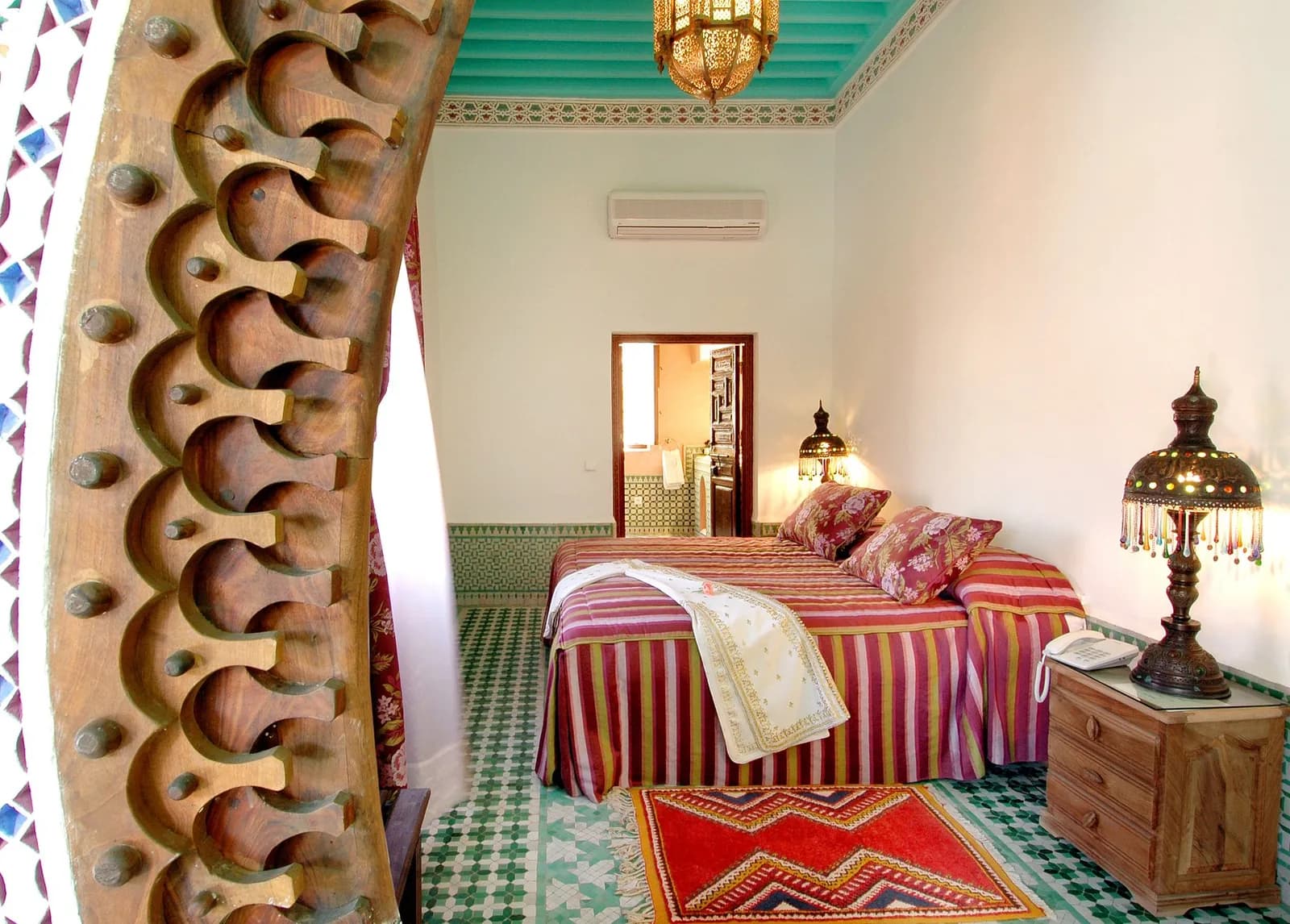 Chambre d'hôtes d'un riad marocain : lit à rayures rouges, sol en zellige vert et blanc, plafond peint turquoise et lanternes en métal ajouré