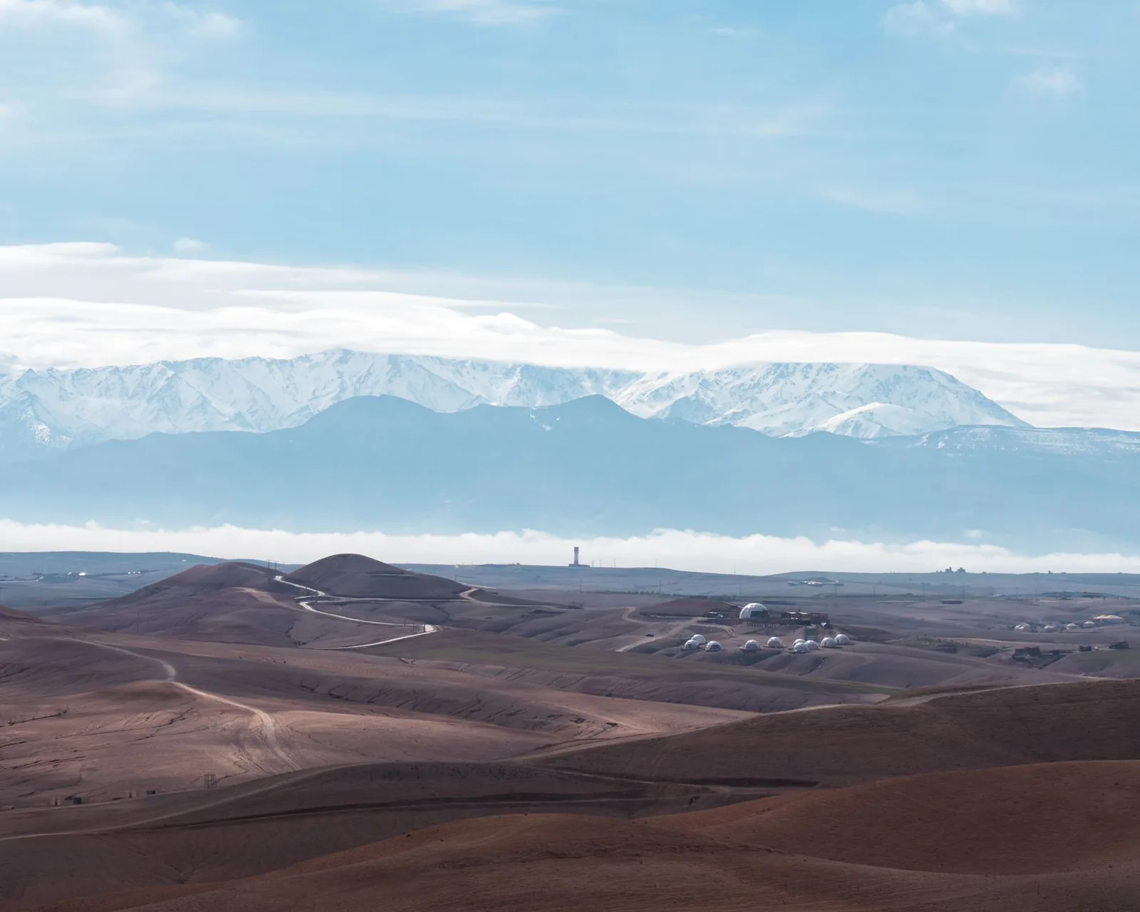 Collines rocailleuses ocre d'un désert avec de petits dômes blancs de camp et la chaîne enneigée de l'Atlas au loin