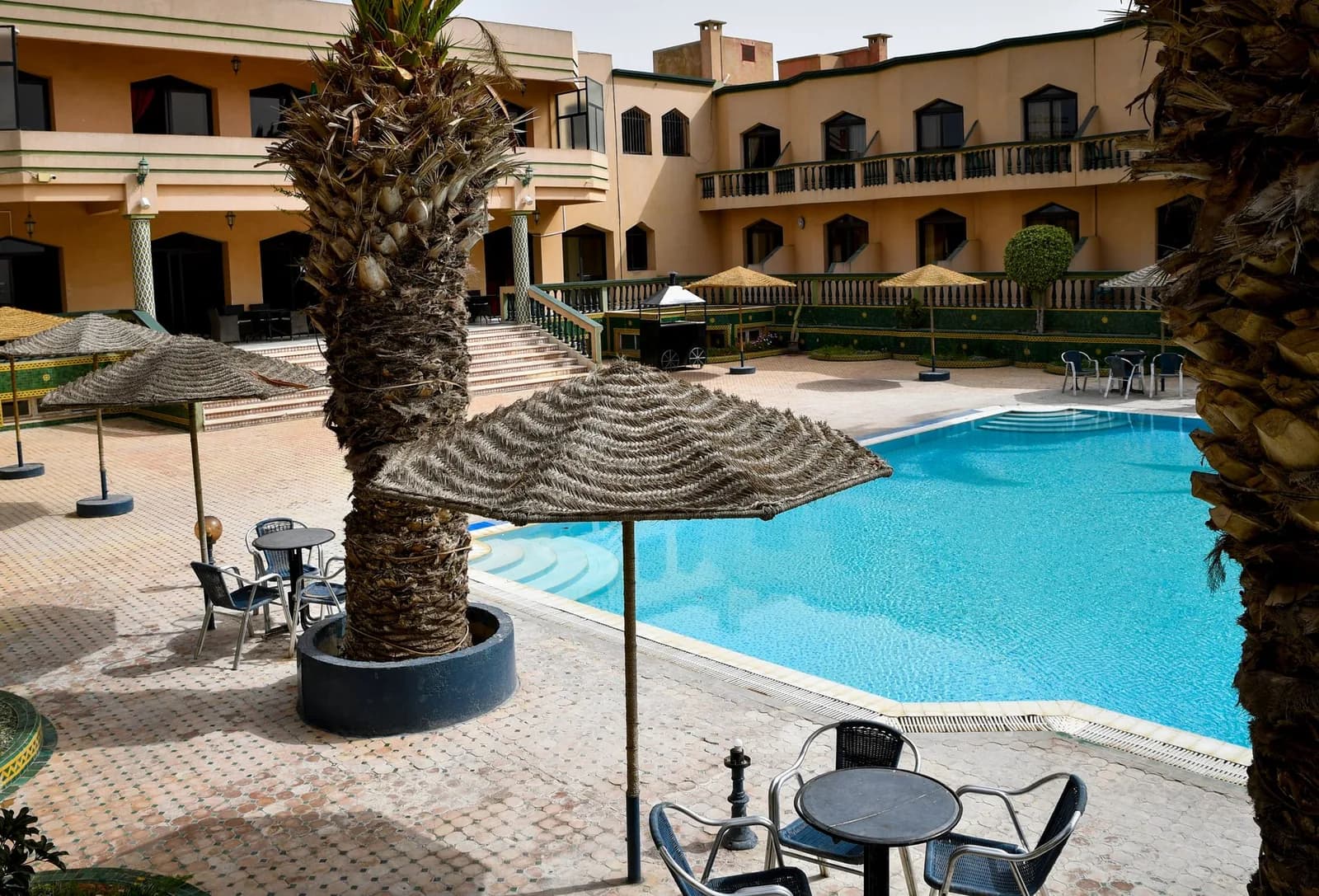Piscine d'hôtel entourée d'une terrasse carrelée, de parasols en paille et de palmiers, sans baigneurs