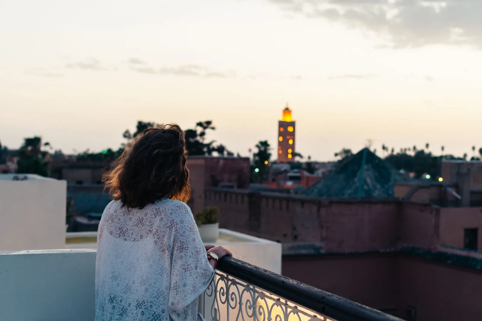 Femme accoudée à un balcon de terrasse au crépuscule, face aux toits de Marrakech et à un minaret éclairé