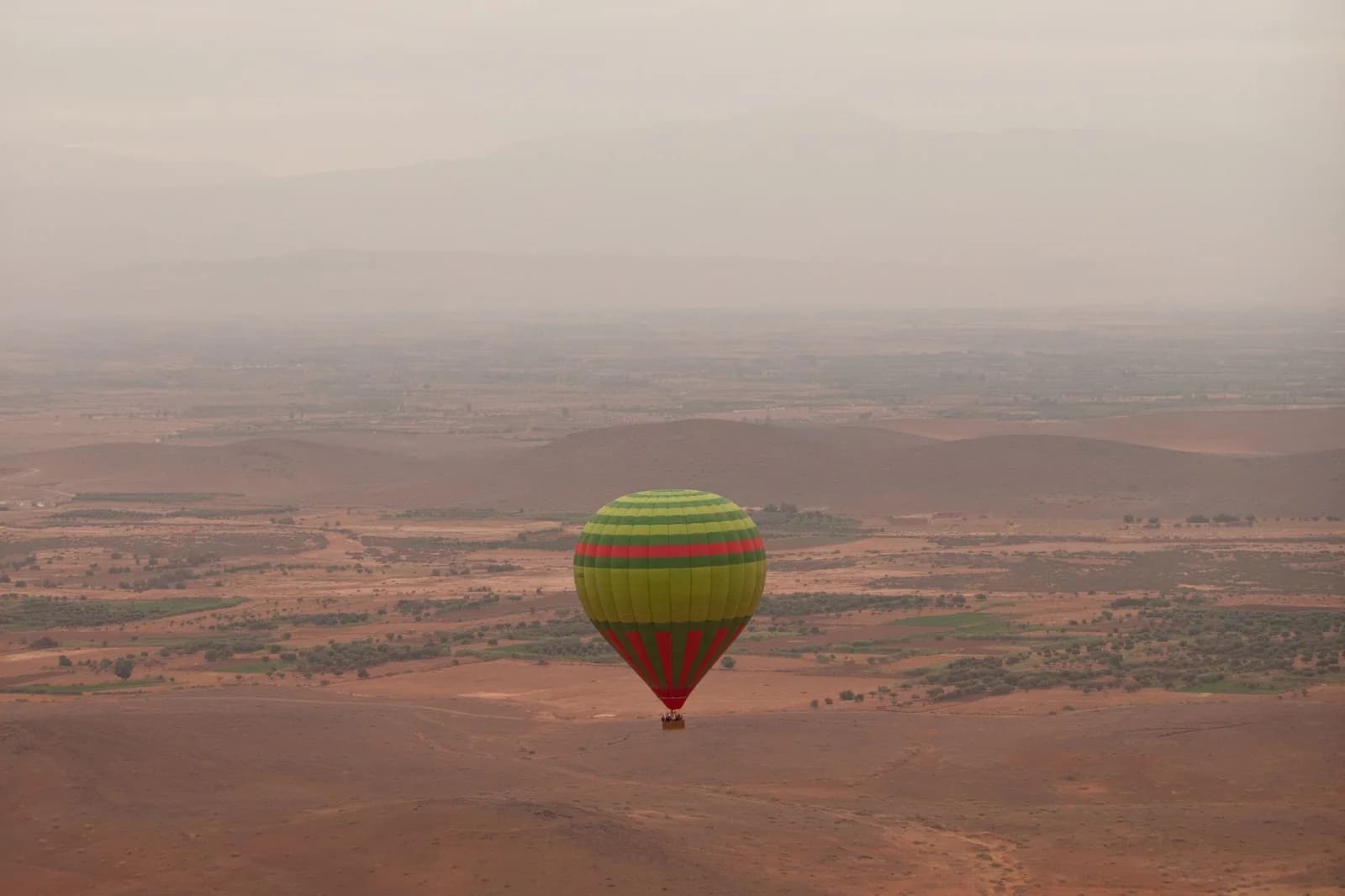 Montgolfière verte et jaune survolant une vaste plaine aride au nord de Marrakech, dans la brume du matin