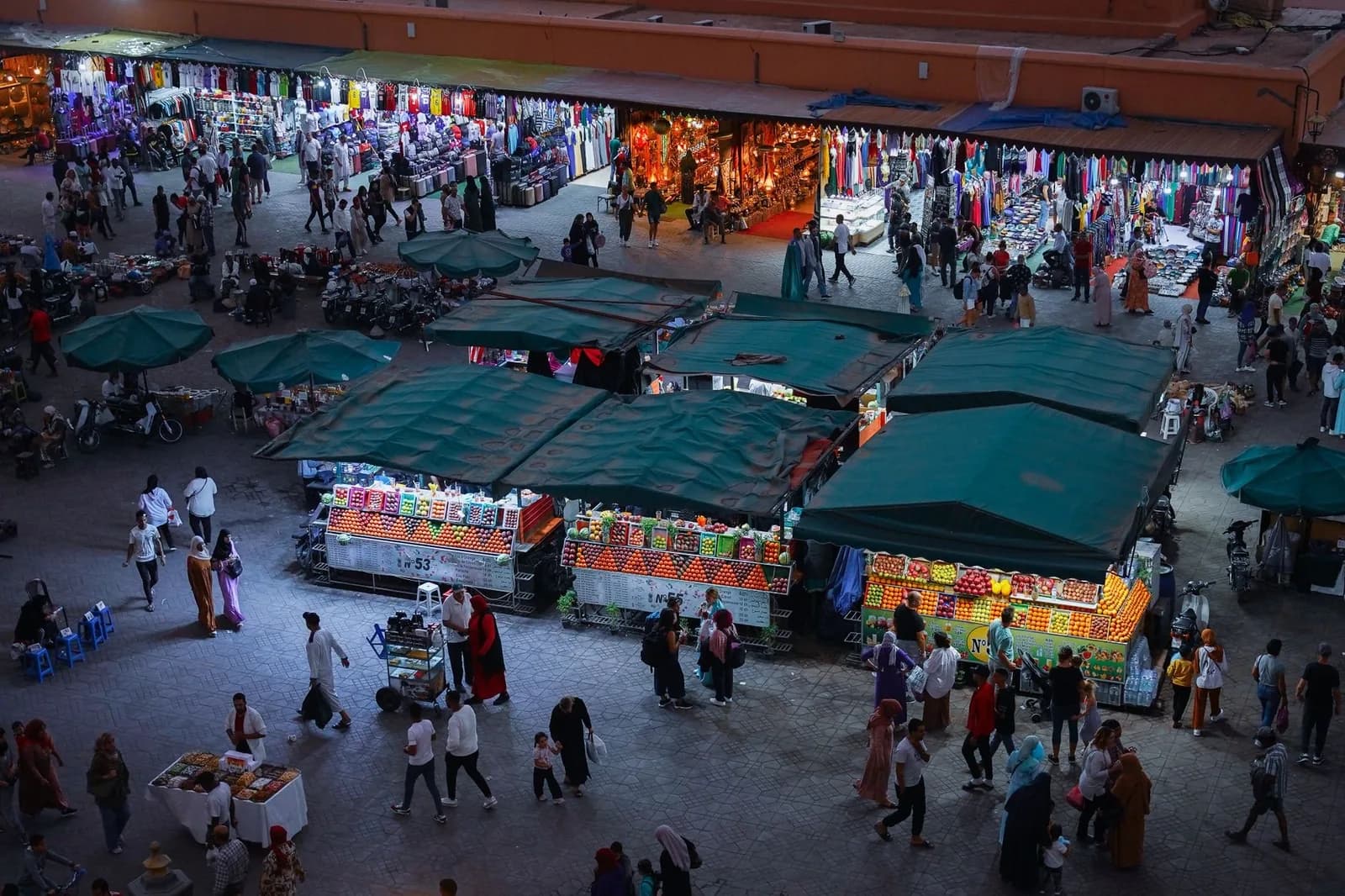 Vue plongeante sur des étals de jus et de fruits sous des auvents verts, dans une grande place de marché animée en soirée