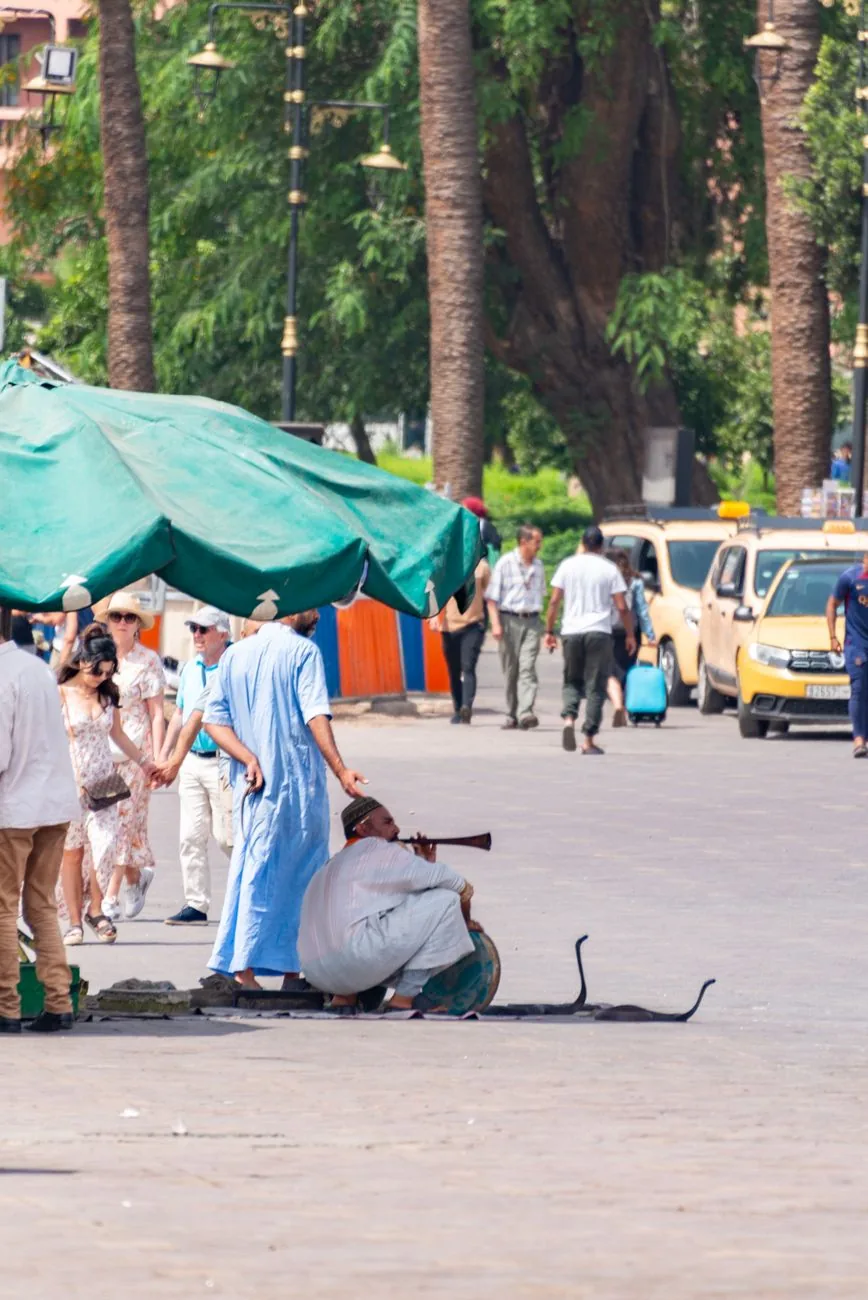 Petits taxis beiges en attente au bord de la place Jemaa el-Fna à Marrakech, avec des passants et un charmeur de serpents