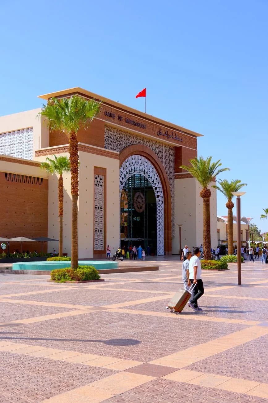 Façade ocre de la gare ONCF de Marrakech avenue Hassan II, avec ses palmiers et des voyageurs tirant leurs valises